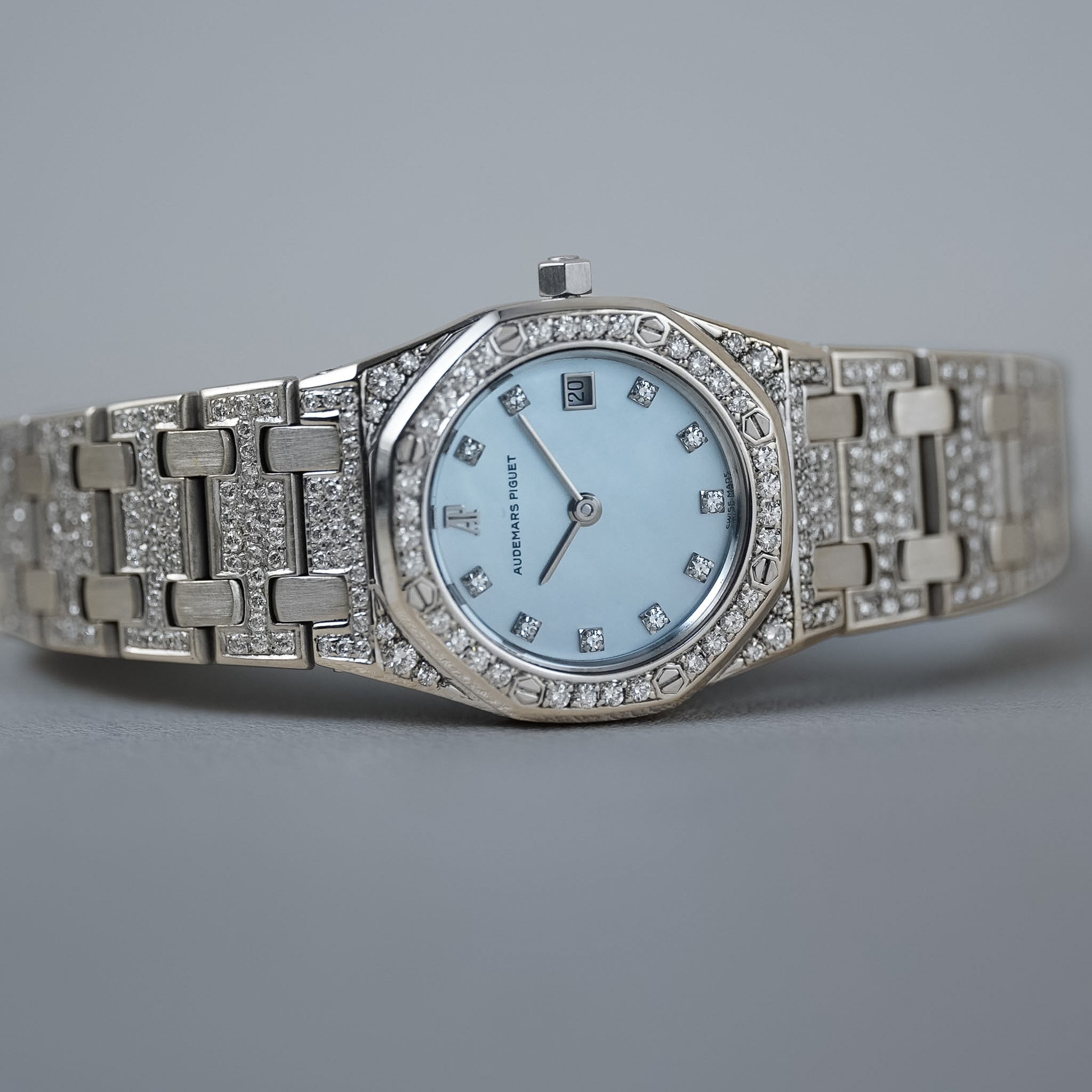 A white gold ladies Audemars Piguet Royal Oak with the case bezel, and bracelet covered in diamonds. The dial is mother of pearl. It's shot laying sideways on a neutral grey background.