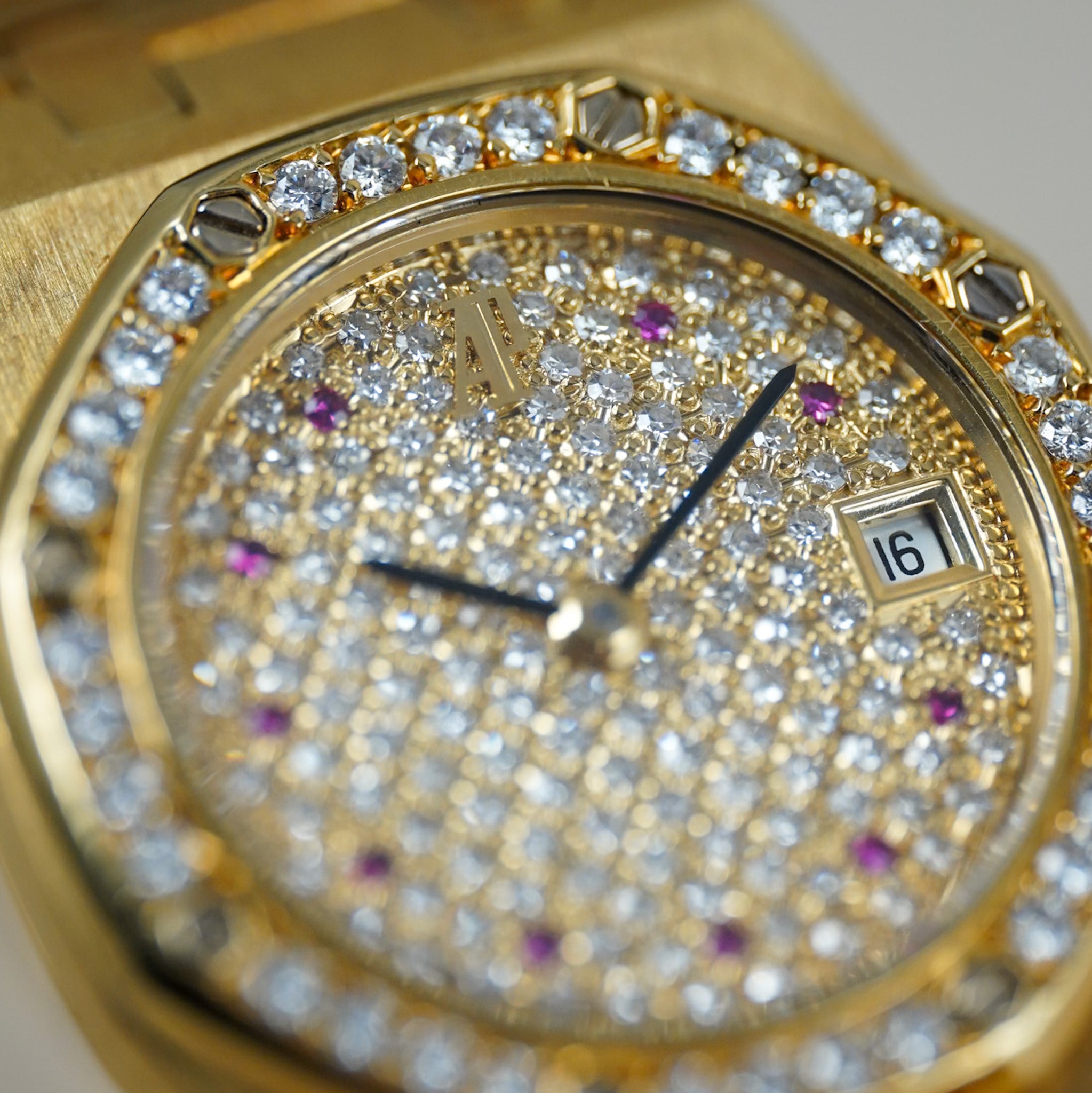 Closeup of a yellow gold ladies Audemars Piguet Royal Oak with a pavé diamond bezel and dial and ruby hour markers, shot on a neutral cream background