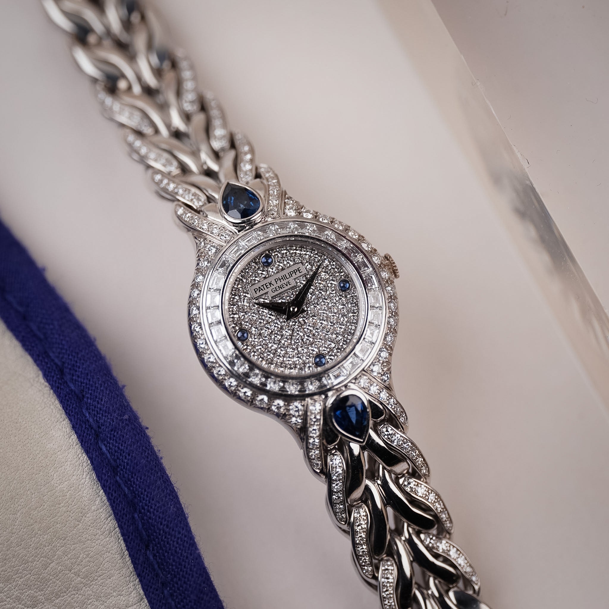 A white gold ladies Patek Philippe watch with a pavé diamond dial, blue sapphire accents, diamond bezel, and a sculpted bracelet. Shot resting flat on a white surface with a blue lined fabric to the left.