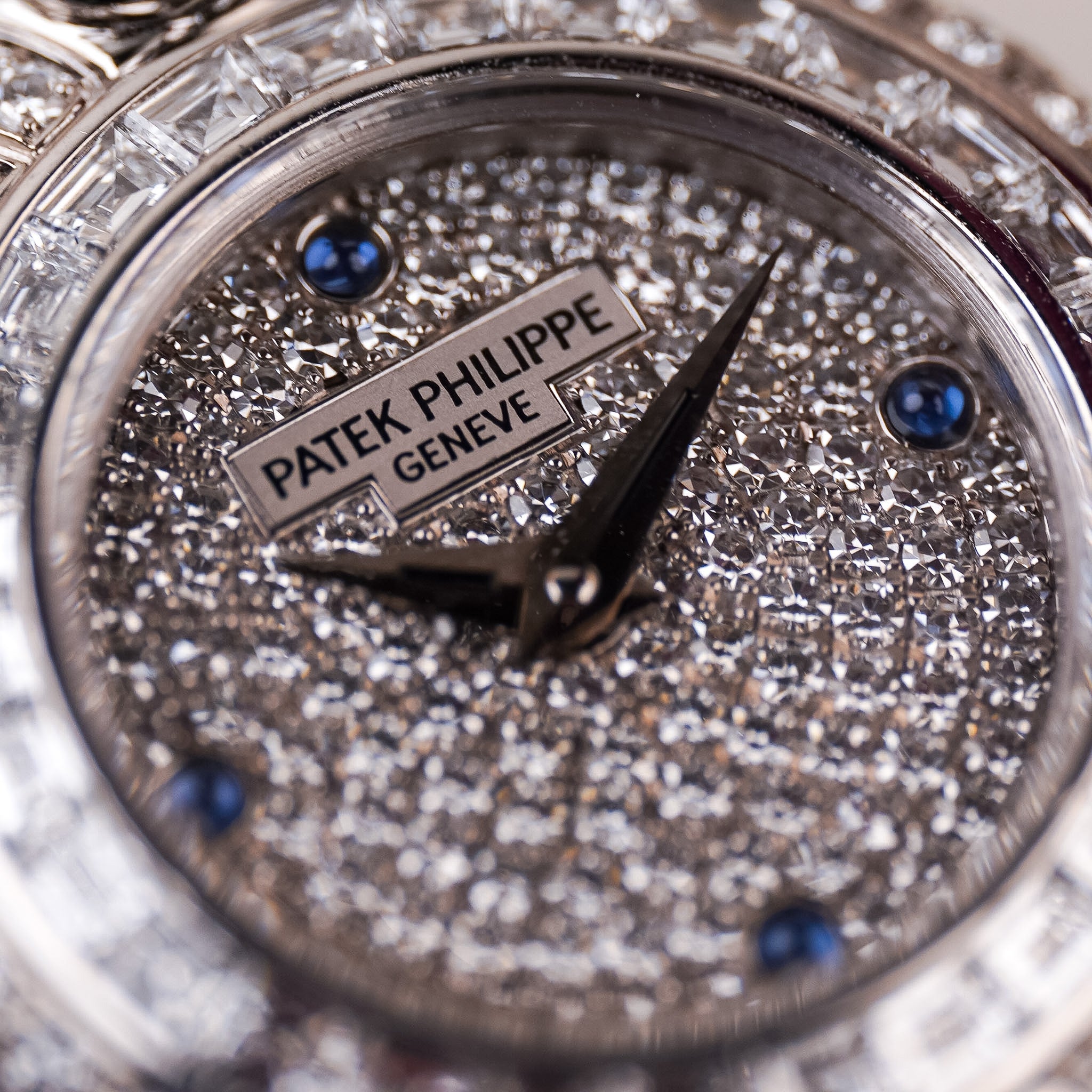 Closeup of a white gold ladies Patek Philippe watch with a pavé diamond dial, blue sapphire accents, and diamond bezel. The Patek Philippe Geneve logo is visible.