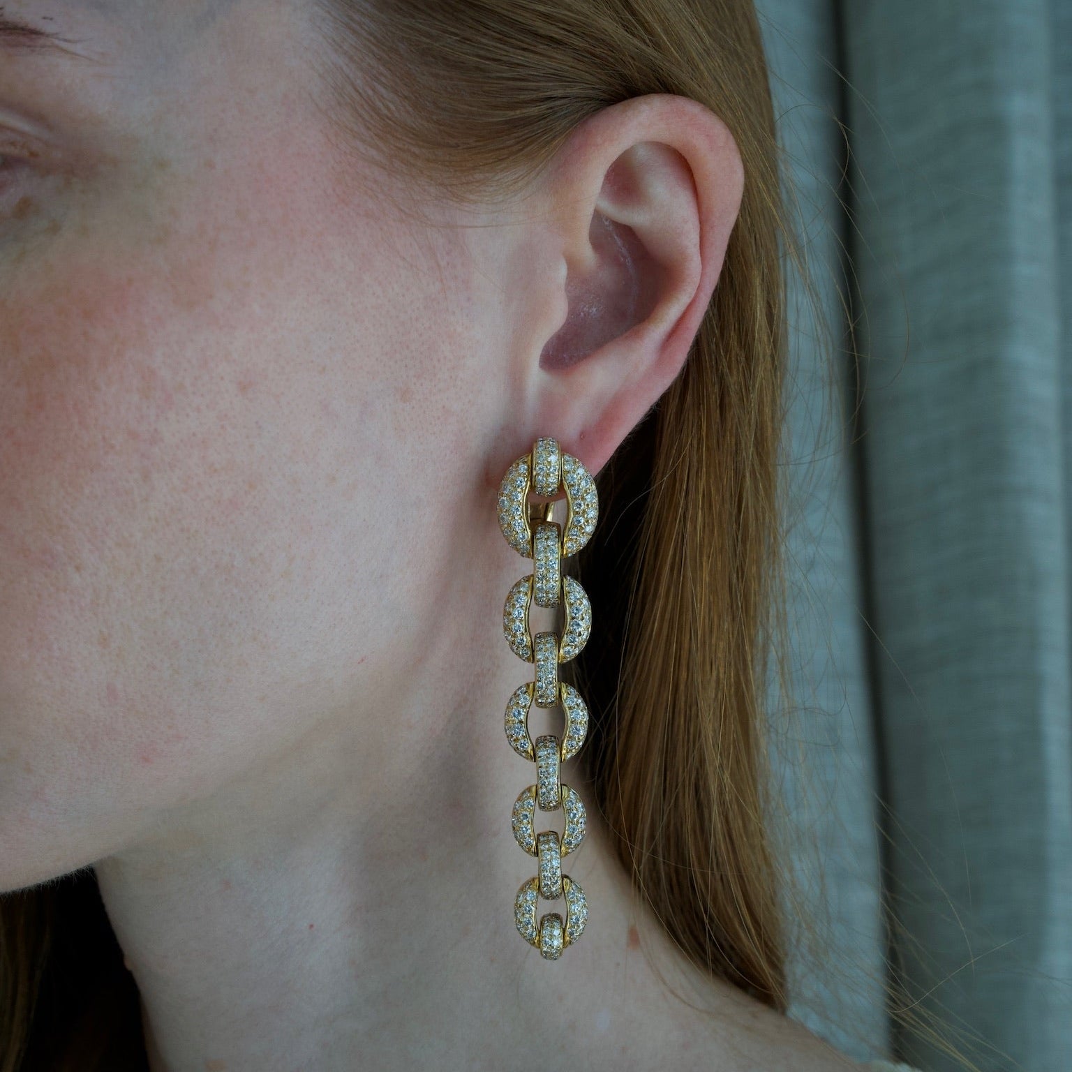 A close-up of a woman's ear wearing a long, dangling link earring with small clear stones set in a gold-tone metal, against a soft gray curtain background.