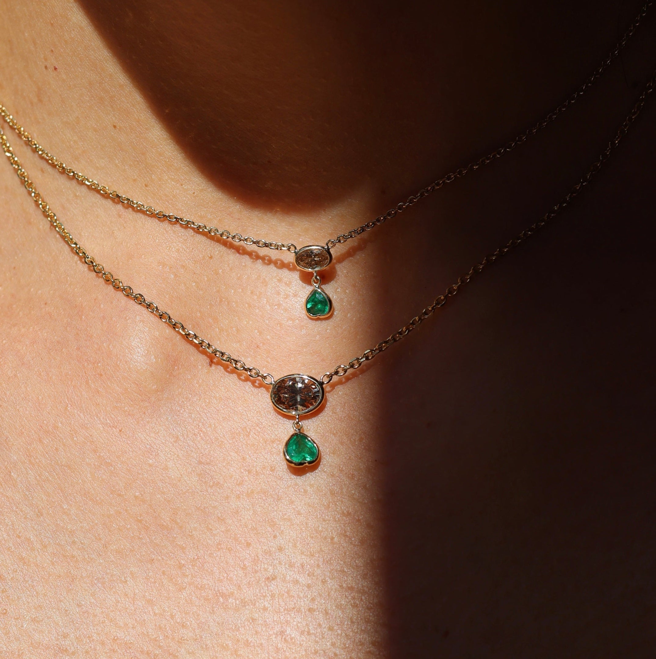 Model wearing two layered 18kt yellow gold necklaces, each with a bezel-set east-west brown oval diamond and a heart-shaped emerald charm, in petite and larger sizes, styled at the collarbone.