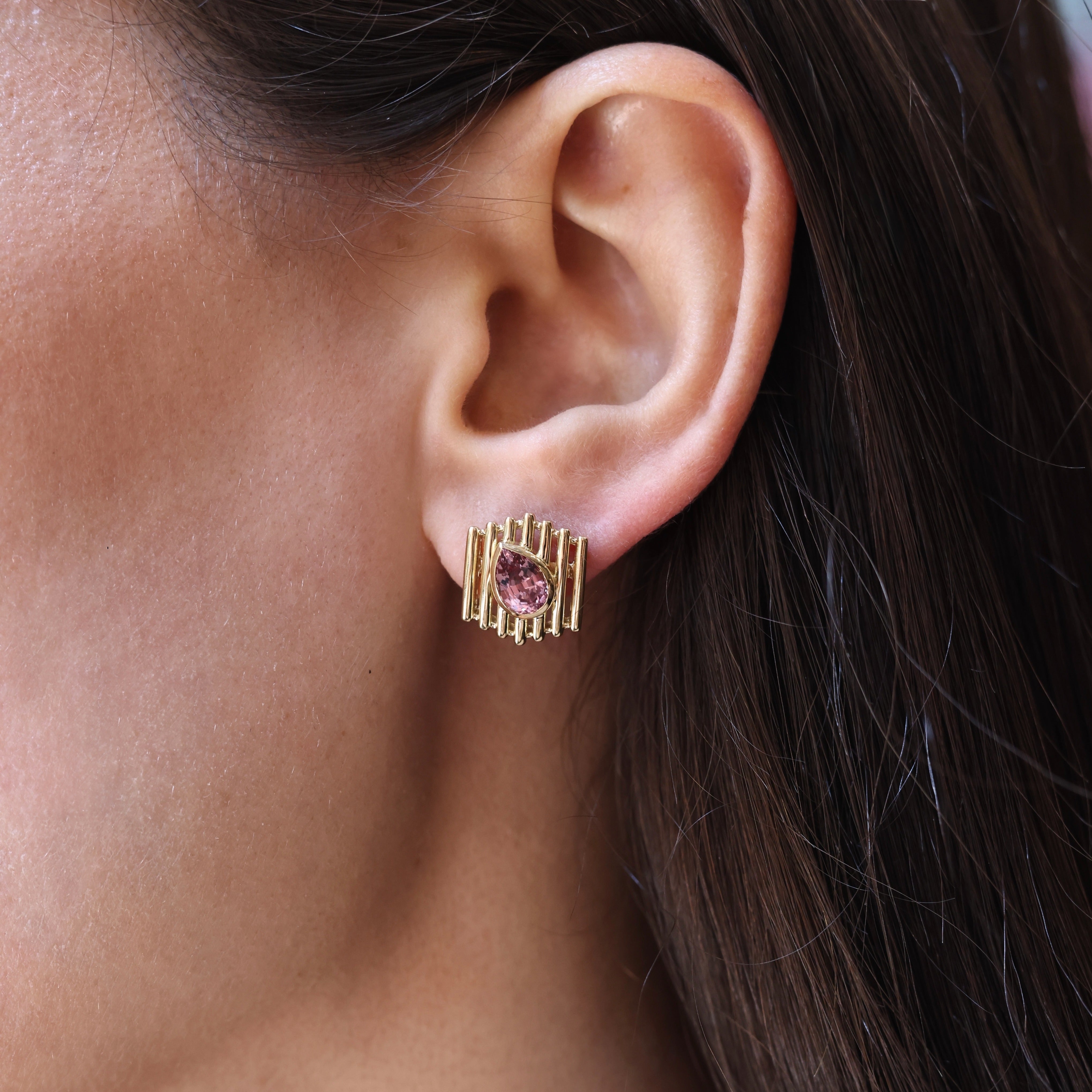 Model wearing a yellow gold cage stud earring with a bezel-set pink pear-shaped gemstone, shown on the earlobe in profile
