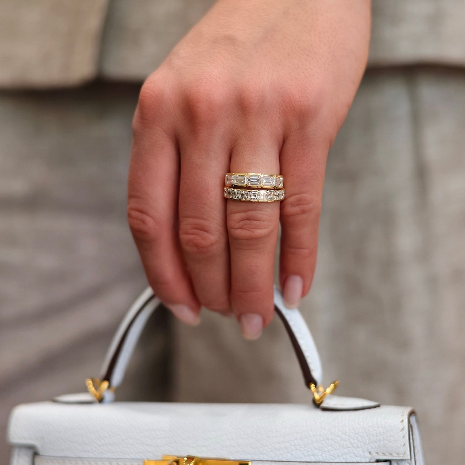 A close-up of a hand holding the top handle of a light gray leather handbag with gold hardware, wearing a double-band ring with clear stones on one finger.