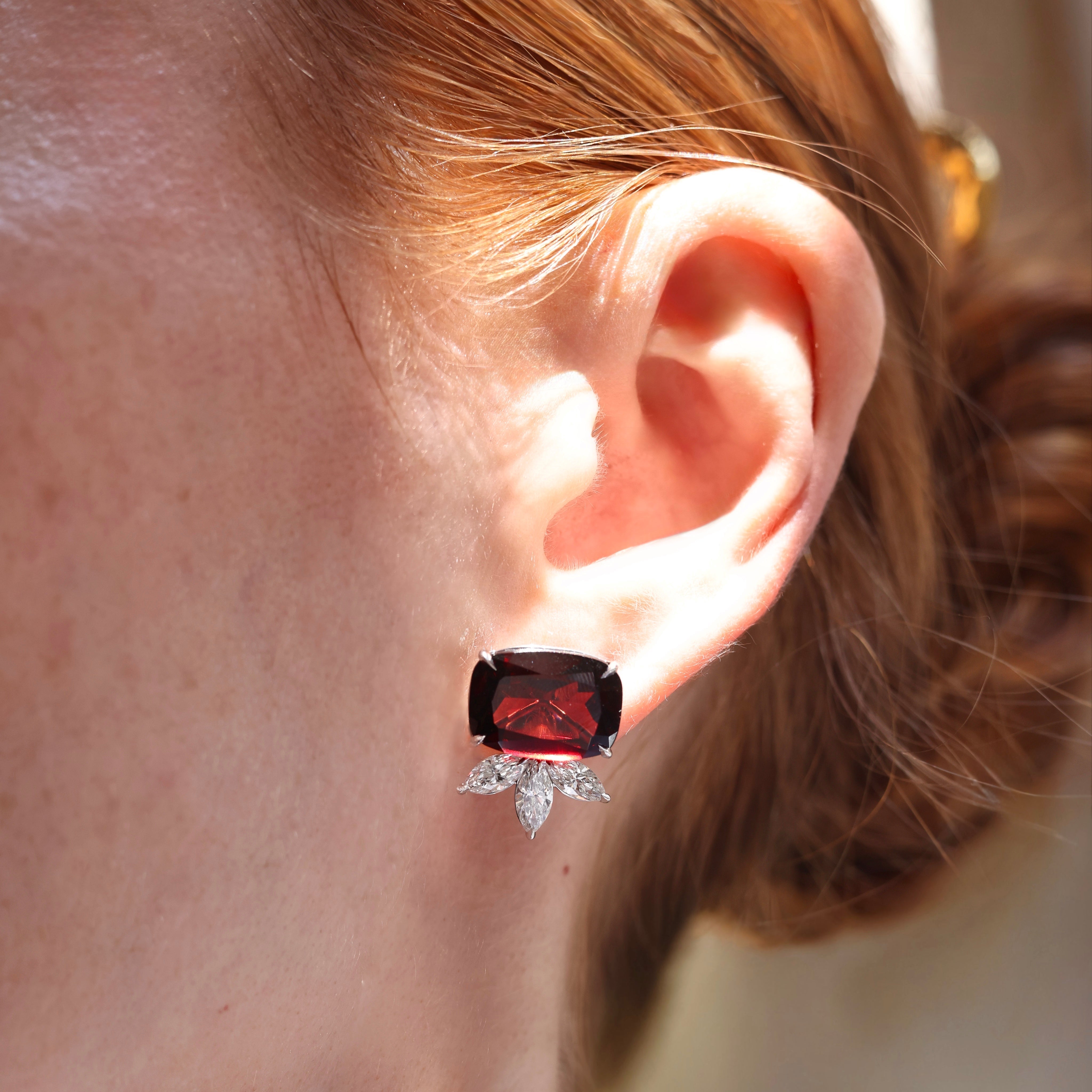 A pair of white gold stud earrings each featuring a large cushion-cut deep red garnet held in four prongs, with a fan of three marquise-cut diamonds set below on model