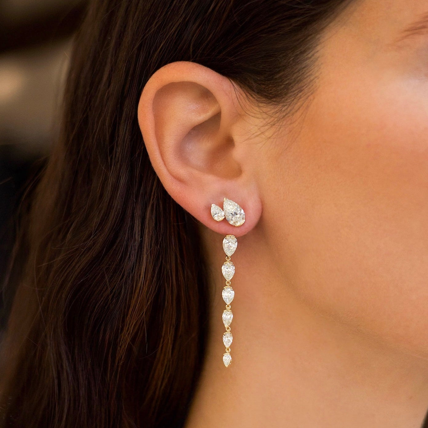 A close-up of a model's ear wearing a dangling statement earring. The earring features a small cluster of diamonds at the top and a vertical drop of pear-shaped diamonds.