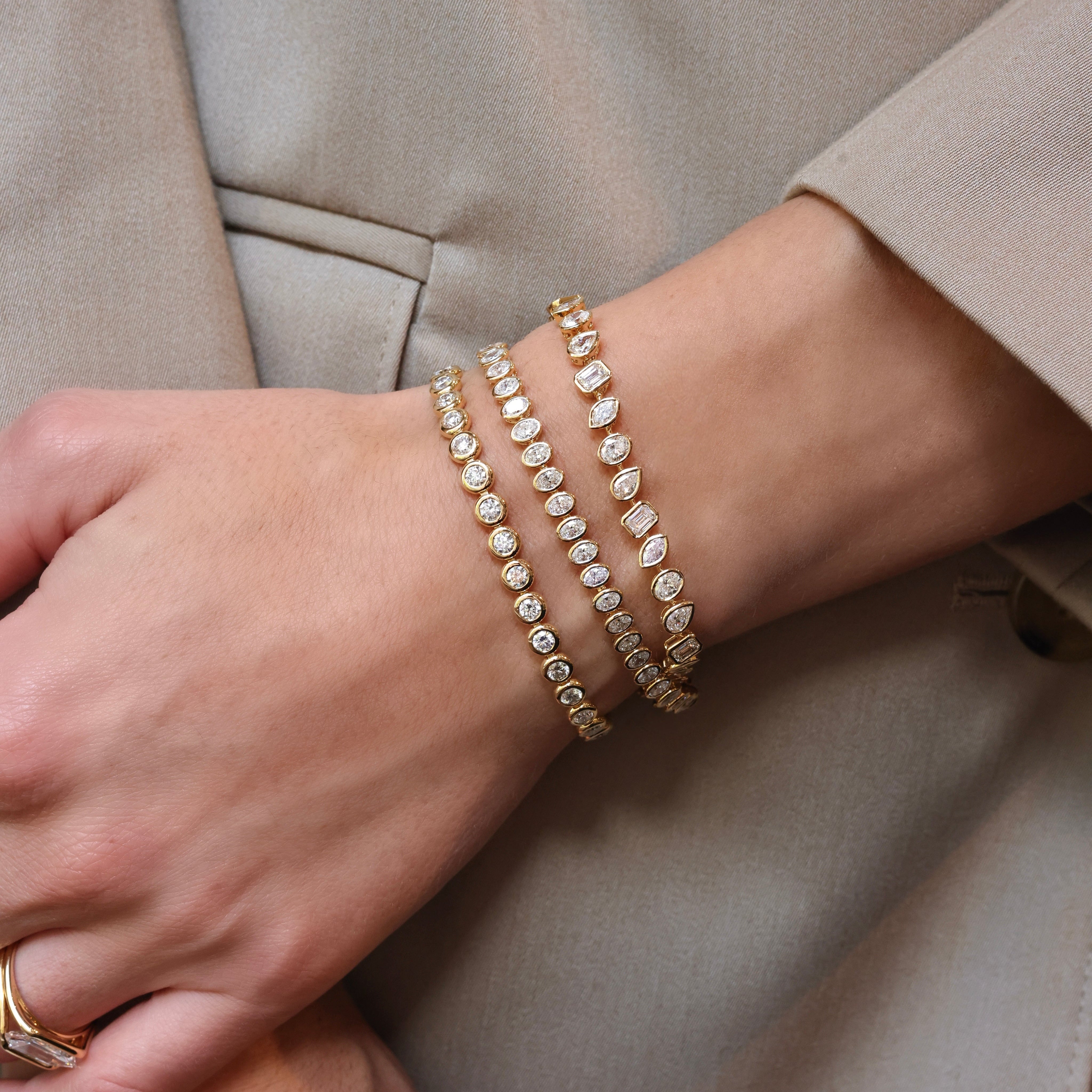 Three bezel-set diamond tennis bracelets in 18K yellow gold, stacked on the wrist over a beige blazer sleeve