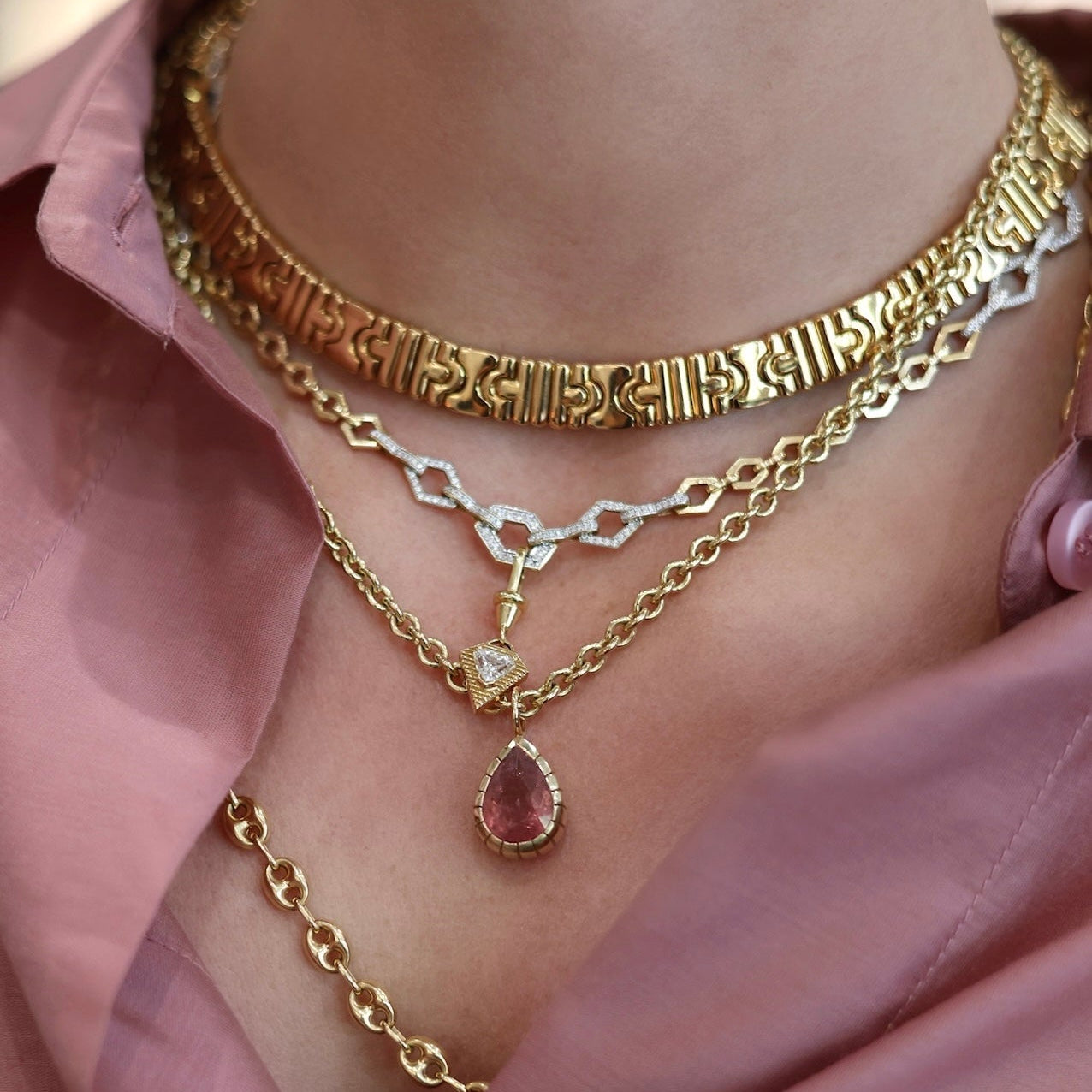 A close-up of a person wearing layered gold necklaces, including a textured choker, a delicate chain necklace with a small heart pendant, and a longer chain with a teardrop pink gemstone pendant, styled with a pink satin shirt.