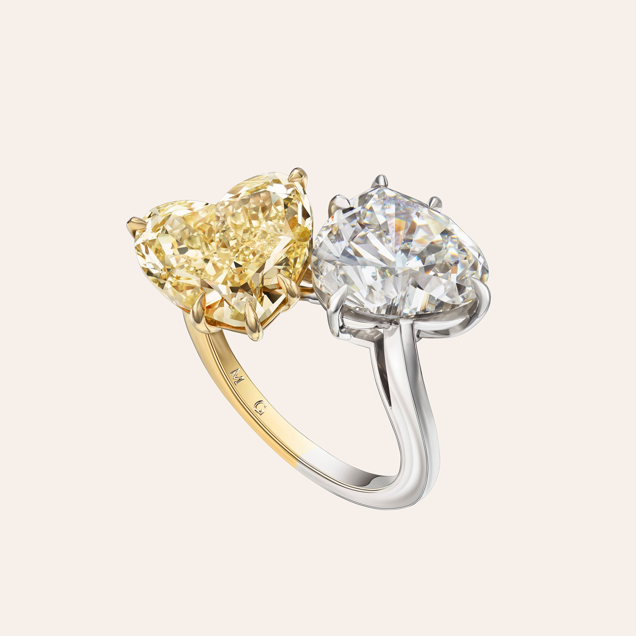 A two-stone diamond ring featuring a yellow heart-shaped diamond and a clear oval diamond, set in a two-tone yellow and white gold band, shown on a light beige background.