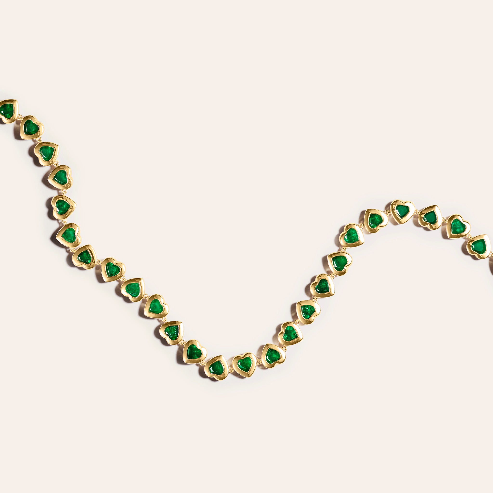 18kt yellow gold anklet composed of bezel-set heart-shaped emeralds in a continuous linked chain, draped in a loose curve against a cream background.