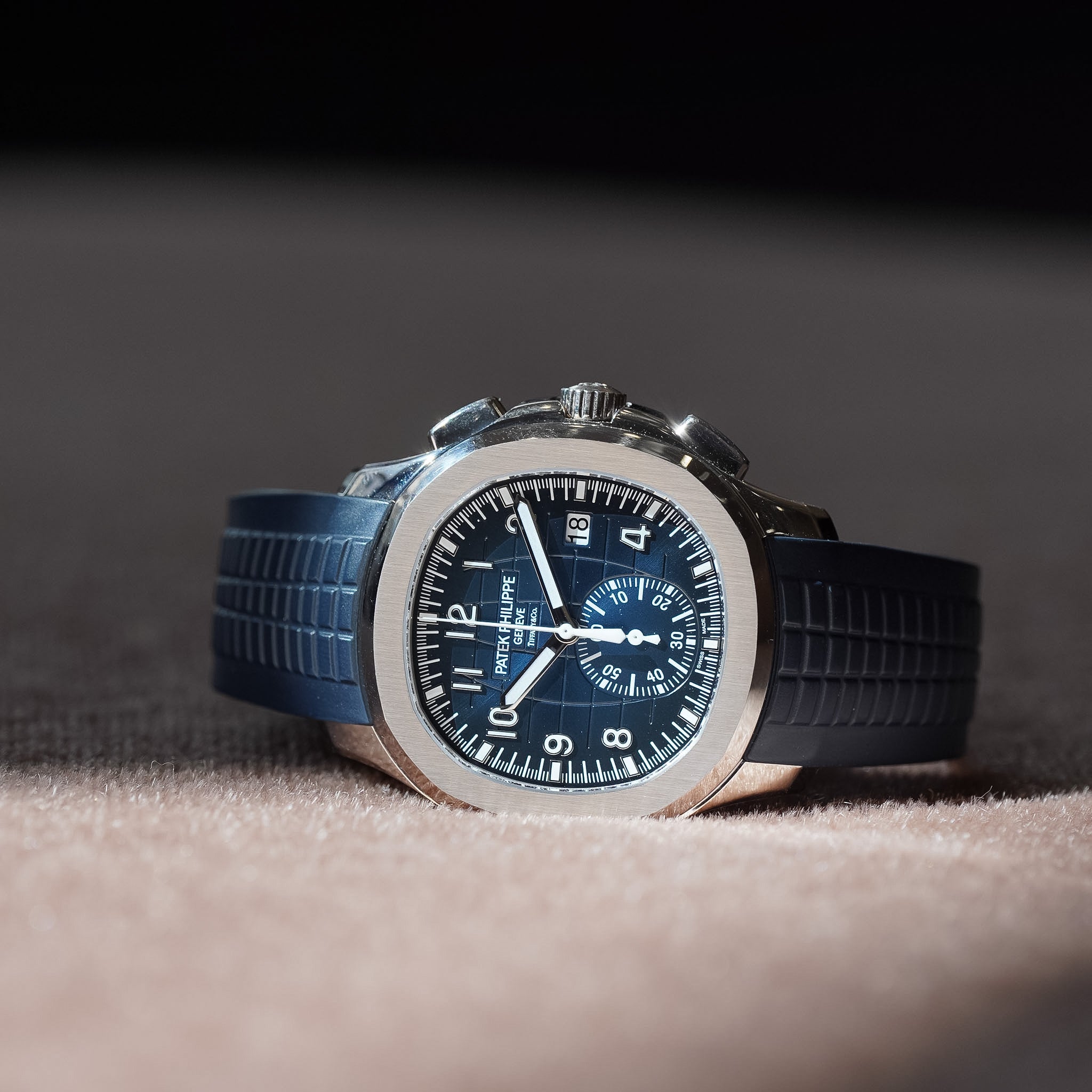 A wristwatch with a silver-toned octagonal bezel, dark blue dial, and dark blue textured strap, photographed on a soft beige surface with a dark background.