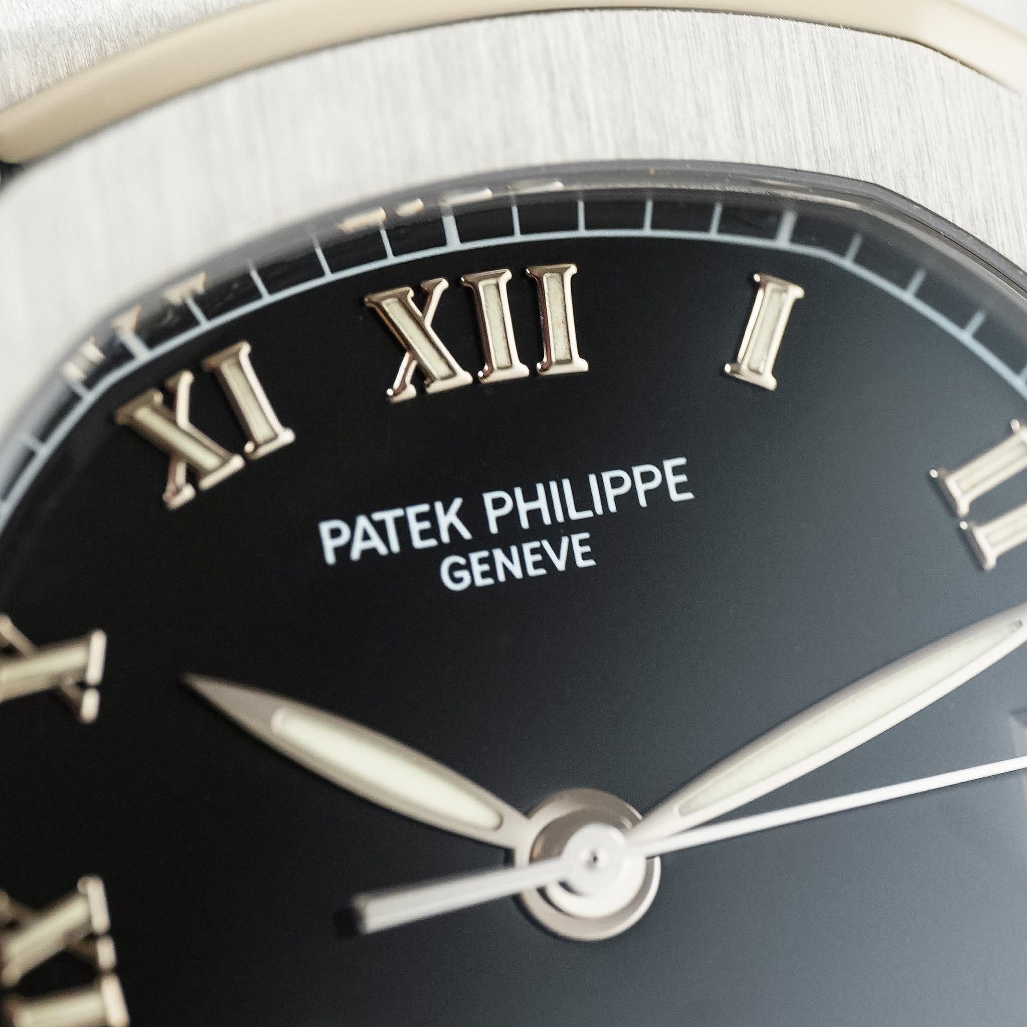 Close-up of a Patek Philippe Geneve watch dial with a black face, silver Roman numerals, silver hands, and a brushed silver-toned case edge.