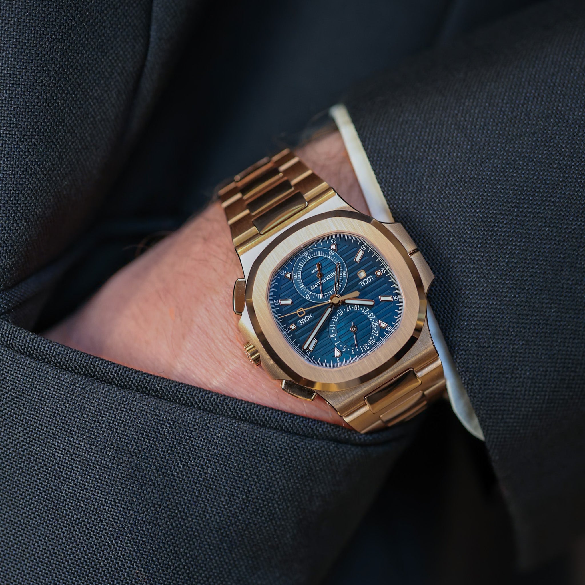 Gold wristwatch with a blue face on a person's wrist, wearing a dark suit.