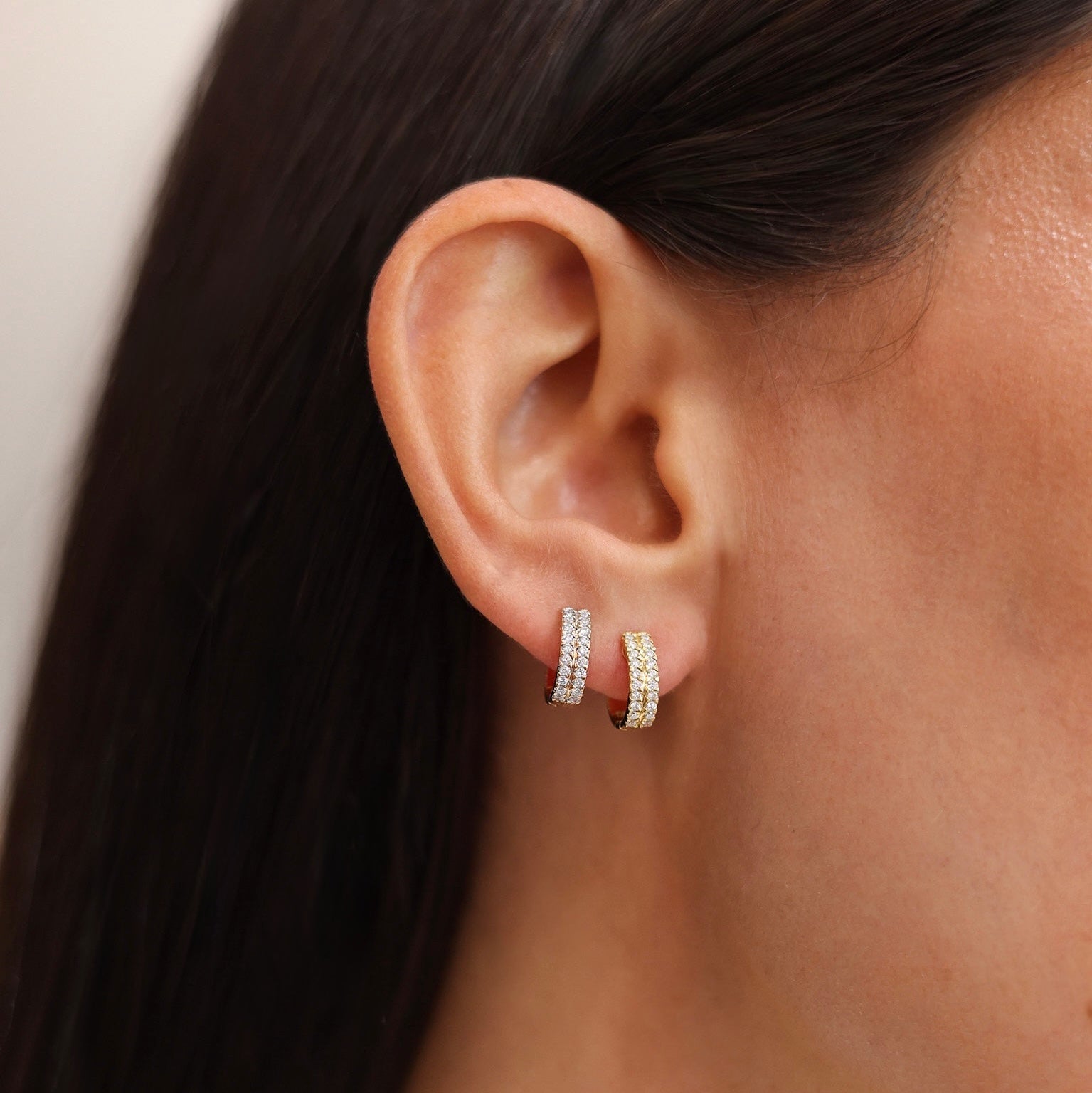 A close-up of a model's ear wearing a pair of small silver hoop earrings with pavé-set clear stones.