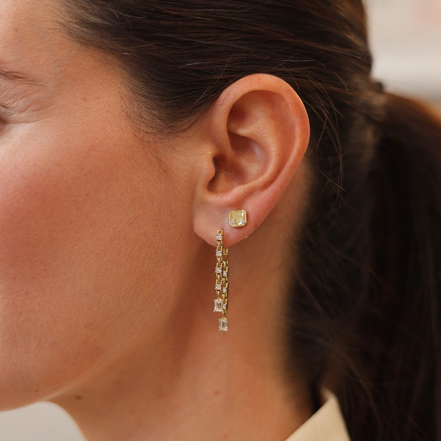 A pair of yellow gold dangle earrings with emerald and round-cut diamonds displayed on a light cream background on model